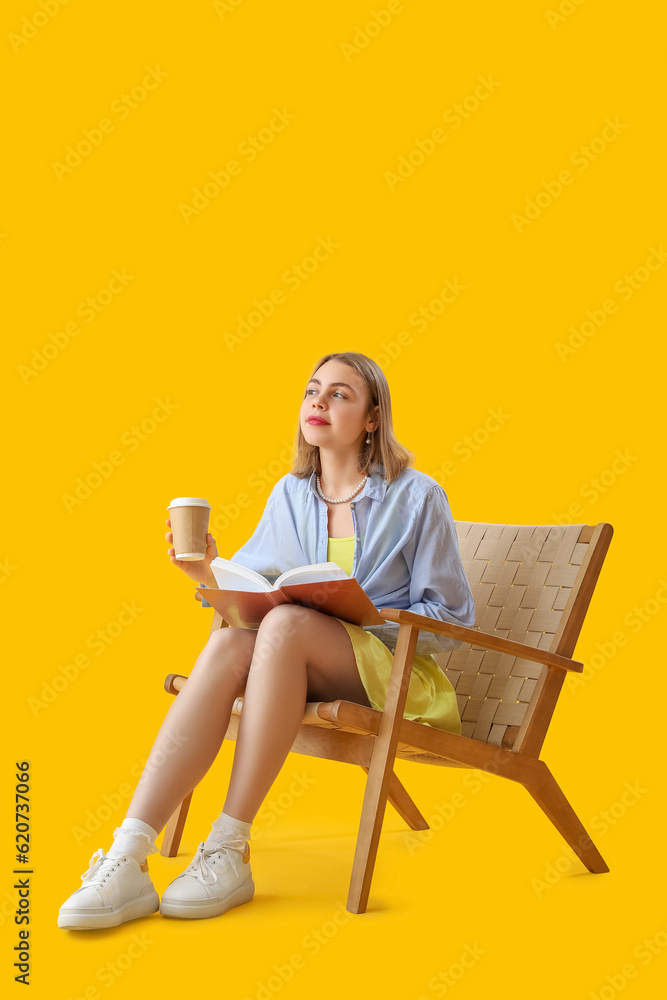 Young woman with cup of coffee and book sitting in wooden armchair on yellow background