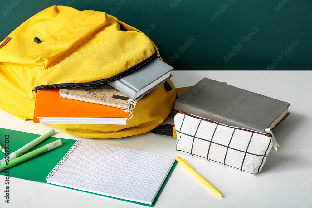 Yellow school backpack with notebooks, pencil case and markers on white wooden table near dark green