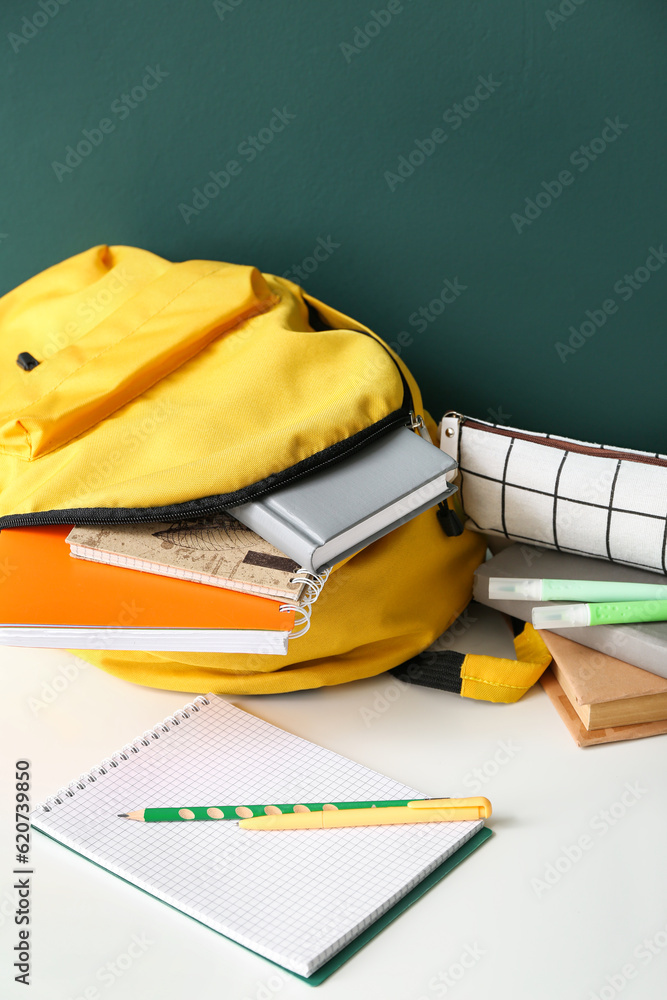 Yellow school backpack with notebooks, pencil case and markers on white wooden table near dark green