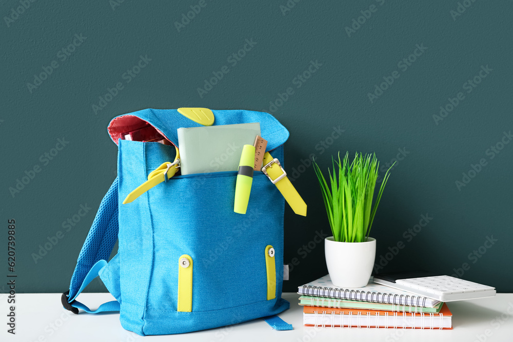 Blue school backpack with notebooks, calculator and houseplant on white wooden table near dark green