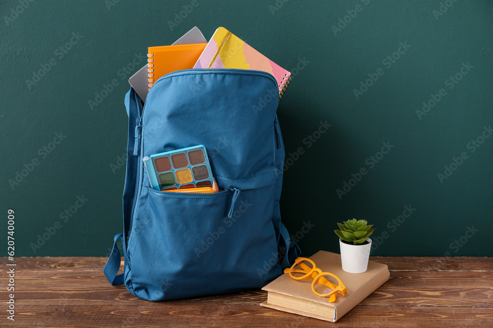 Blue school backpack with notebooks, eyeglasses and succulent on brown wooden table near dark wall