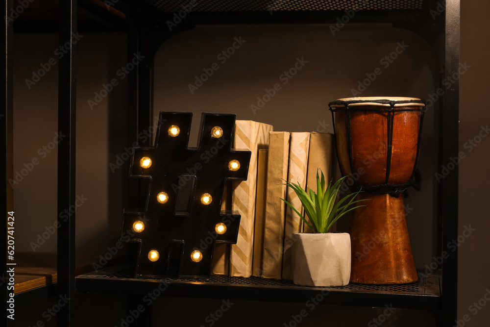 Glowing lamp with books, plant and drum on shelf in dark room