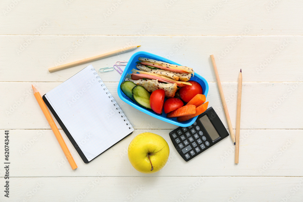 Stationery and lunch box with tasty food on white wooden background