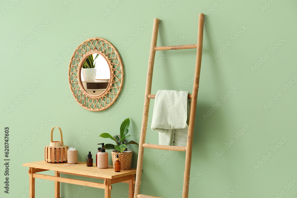 Wooden ladder, table with cosmetic products and mirror hanging on green wall in room