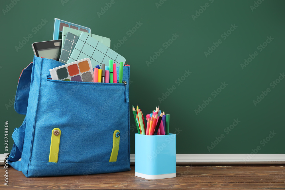 Blue school backpack with notebooks, watercolors and pencils on wooden table near green chalkboard