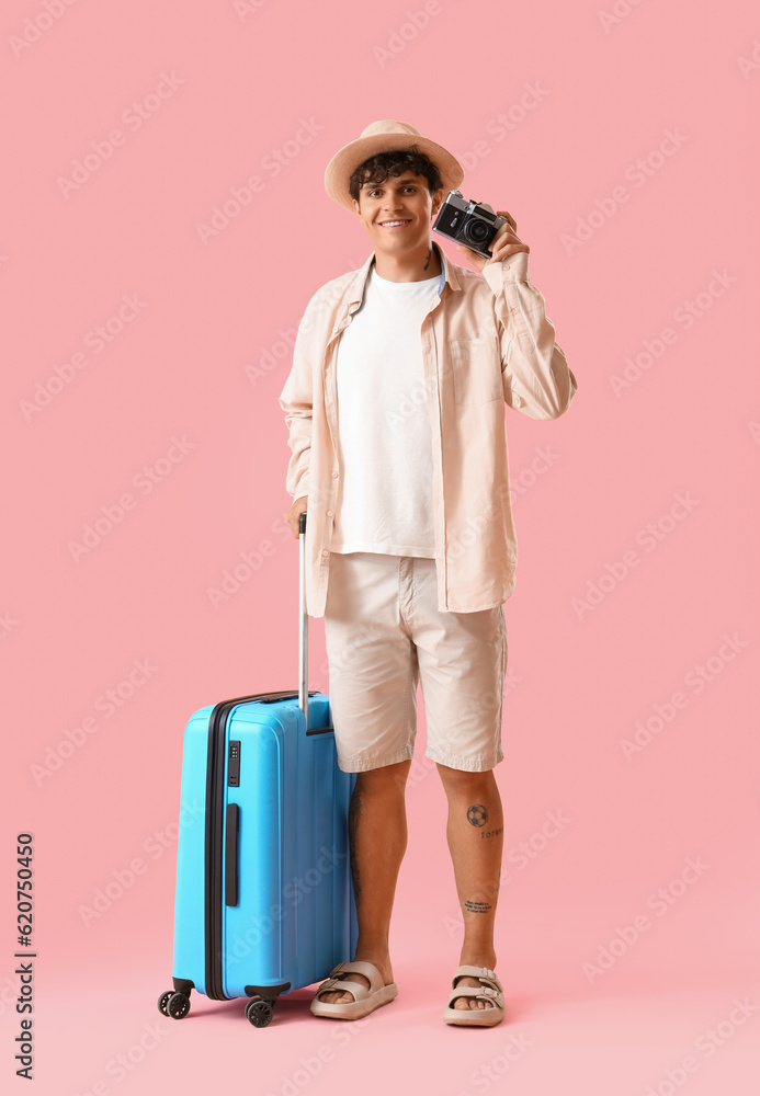 Young man with photo camera and suitcase on pink background