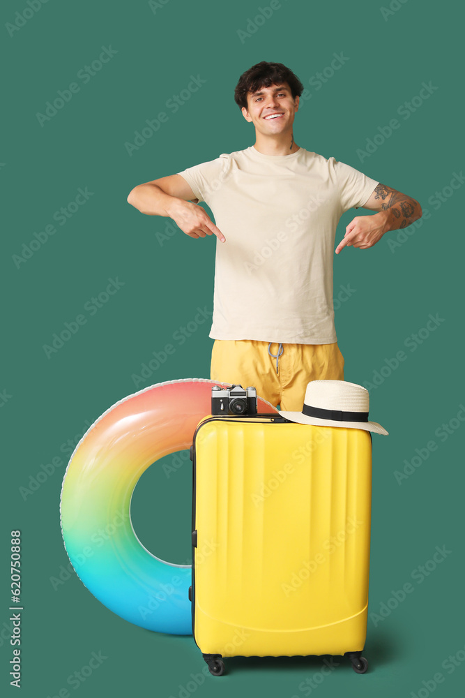 Young man pointing at suitcase with camera and hat on green background