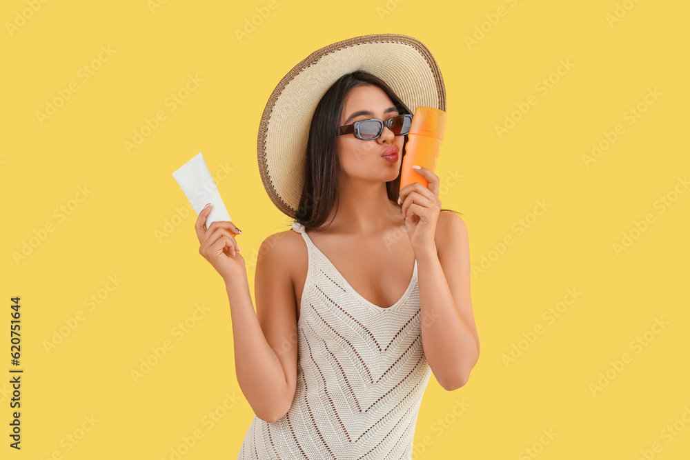 Beautiful young woman with sunscreen cream blowing kiss on yellow background