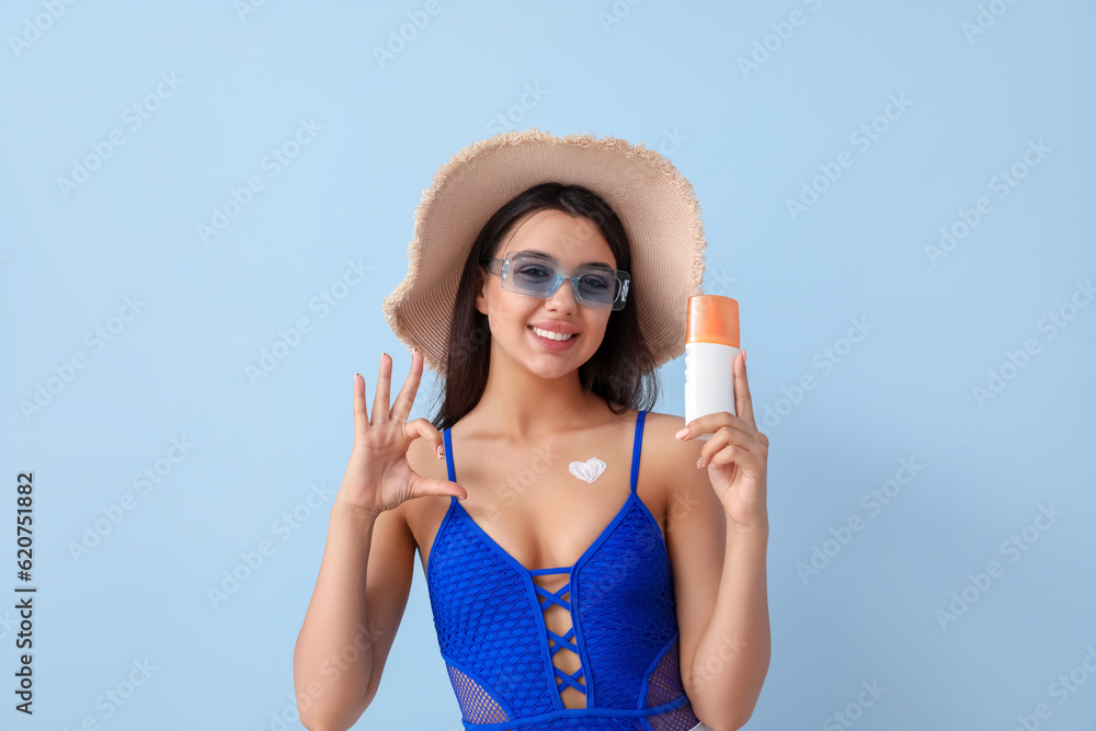 Beautiful young woman with sunscreen cream making heart gesture on light background