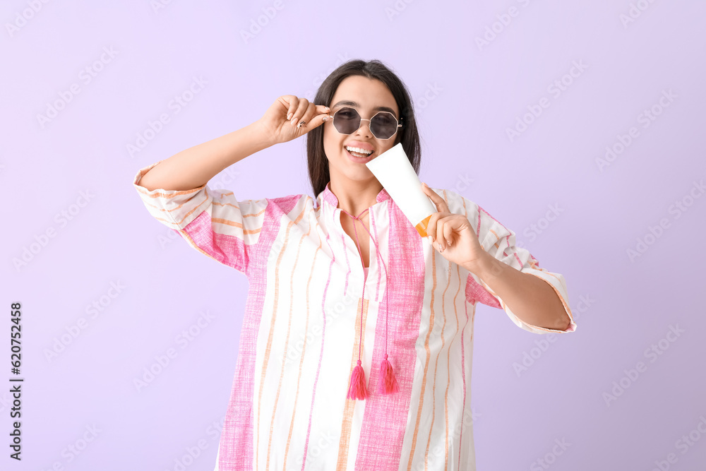 Beautiful young woman with sunscreen cream on lilac background