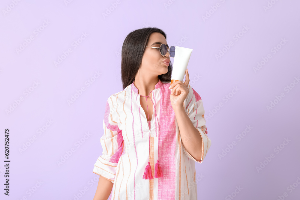 Beautiful young woman with sunscreen cream blowing kiss on lilac background