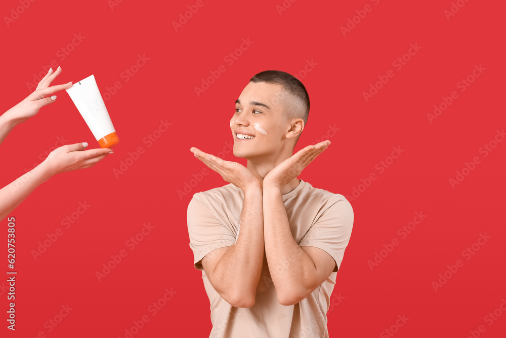 Happy young man and hands holding bottle of sunscreen cream on red background