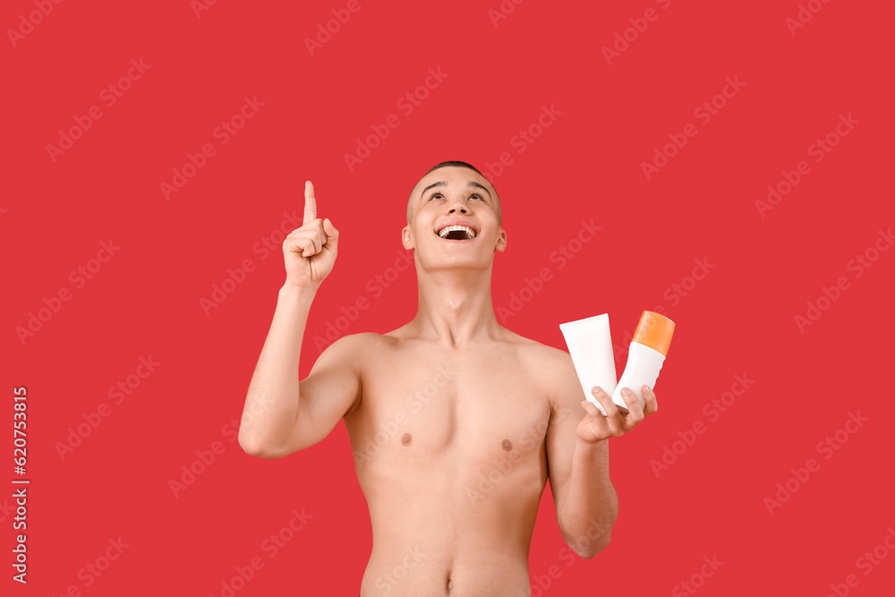 Happy young man with bottles of sunscreen cream pointing at something on red background