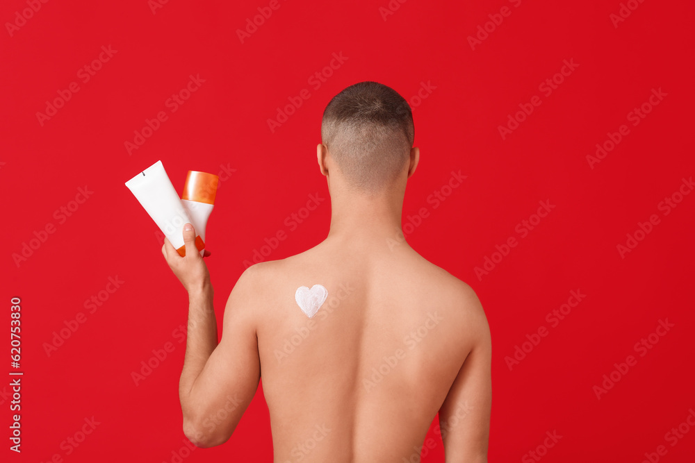 Young man with bottles of sunscreen cream on red background, back view