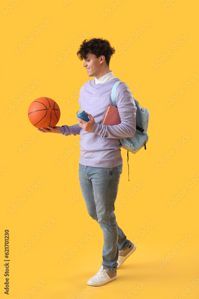 Male student with ball, soda and book on yellow background