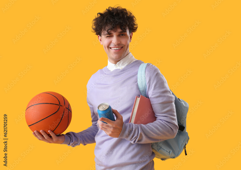 Male student with ball, soda and book on yellow background