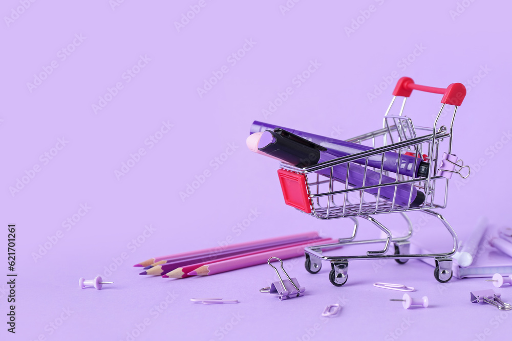 Shopping cart with different stationery on lilac background