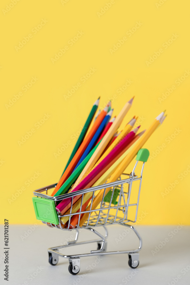 Shopping cart with color pencils on table against yellow background