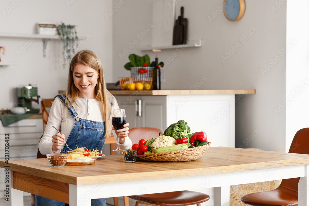 Young woman with glass of wine eating tasty pasta in kitchen