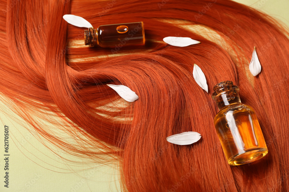 Composition with ginger hair strand, bottles of essential oil and flower petals on green background,