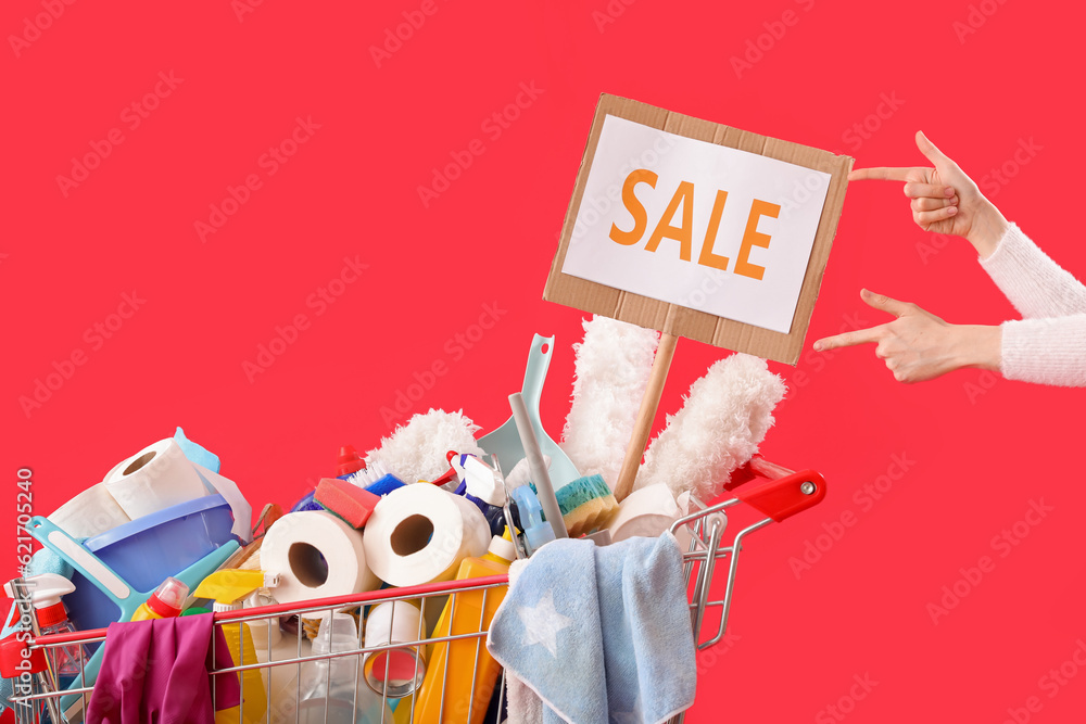 Woman with shopping cart full of cleaning supplies and word SALE on red background