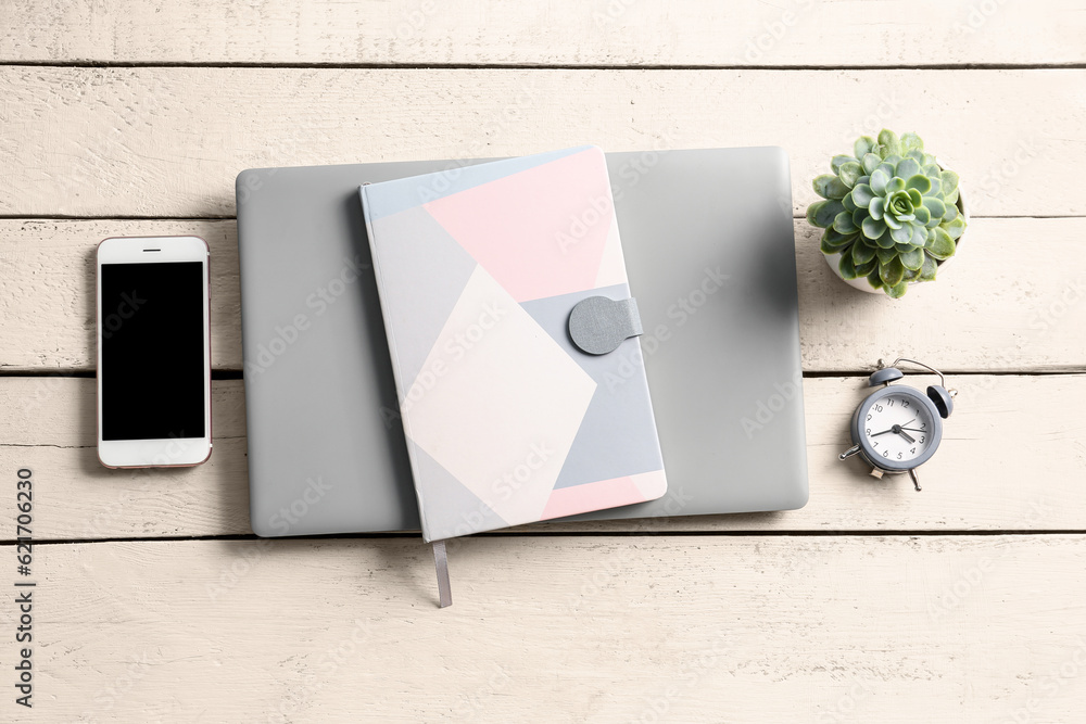 Notebook, modern laptop, mobile phone and alarm clock on white wooden background