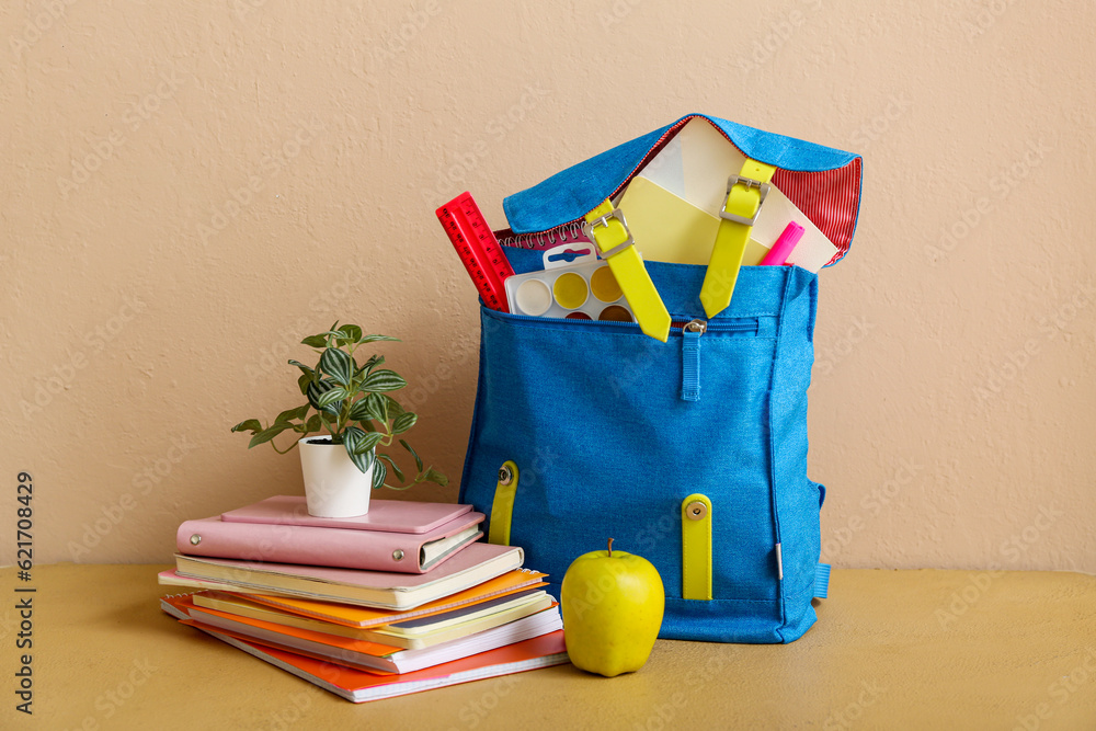 Blue school backpack with notebooks, houseplant and apple on table near beige wall