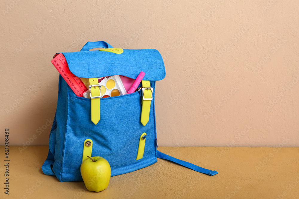 Blue school backpack with apple, watercolor and ruler on table near beige wall