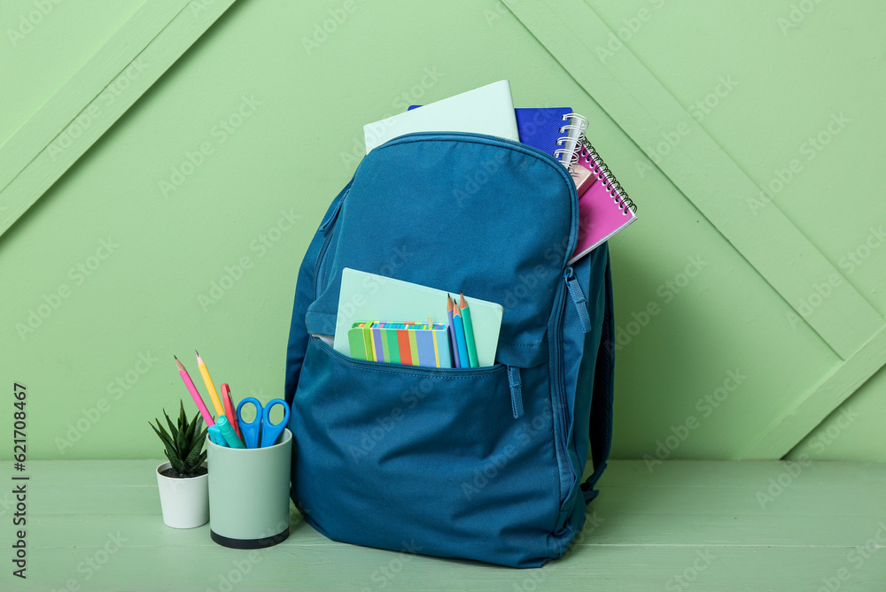 Blue school backpack with notebooks, pencils and houseplant on wooden table near green wall