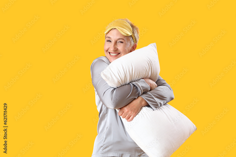 Mature woman in pajamas with pillow on yellow background