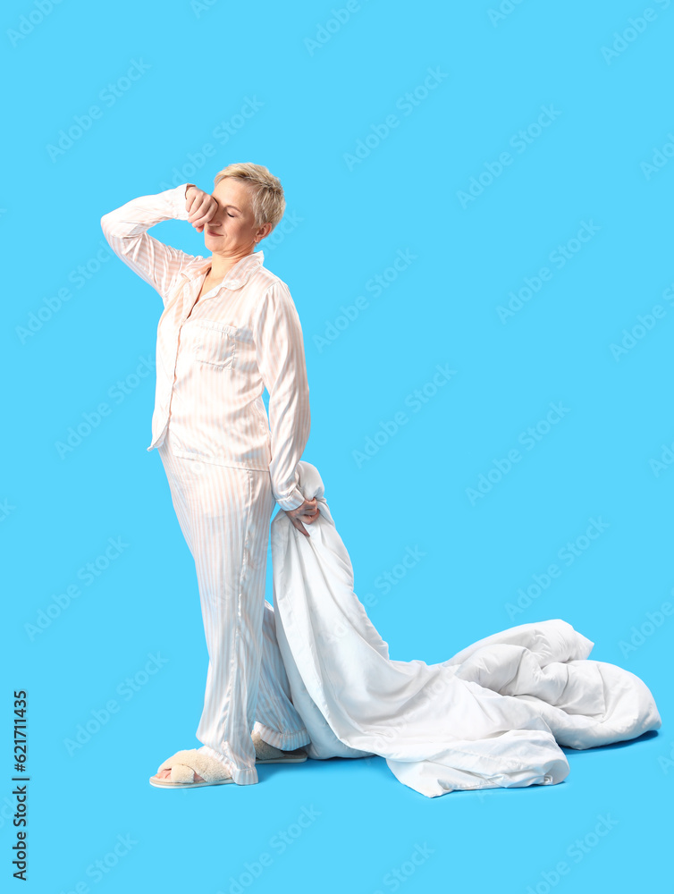 Sleepy mature woman in pajamas with blanket on blue background