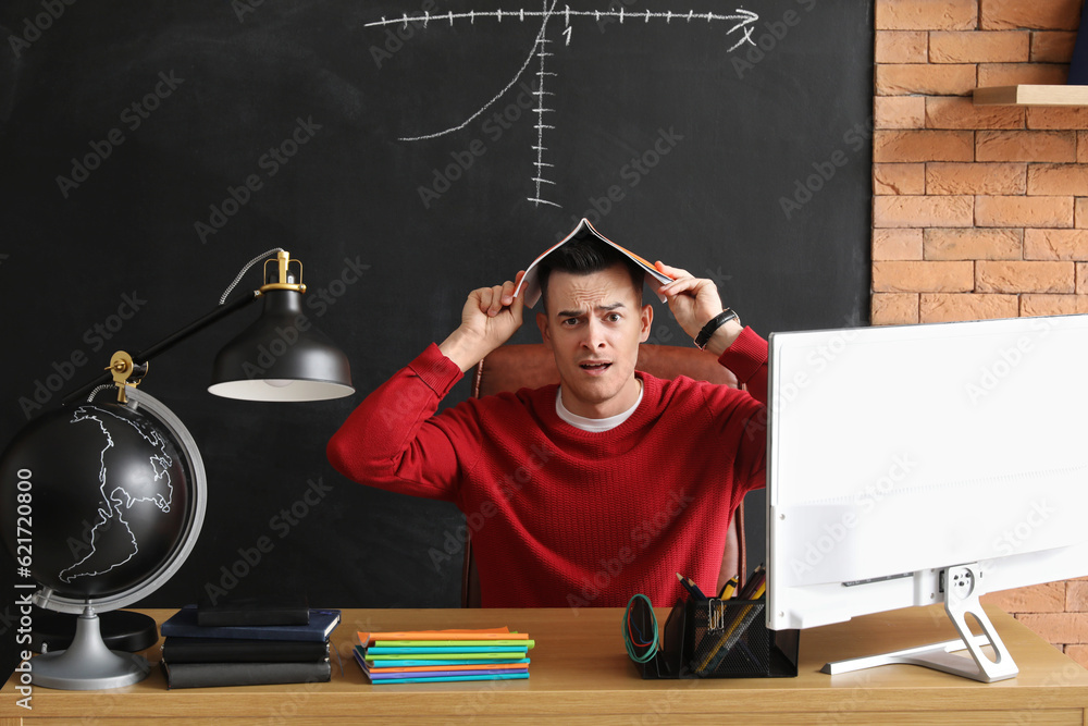 Stressed male teacher with copybook sitting at table in classroom