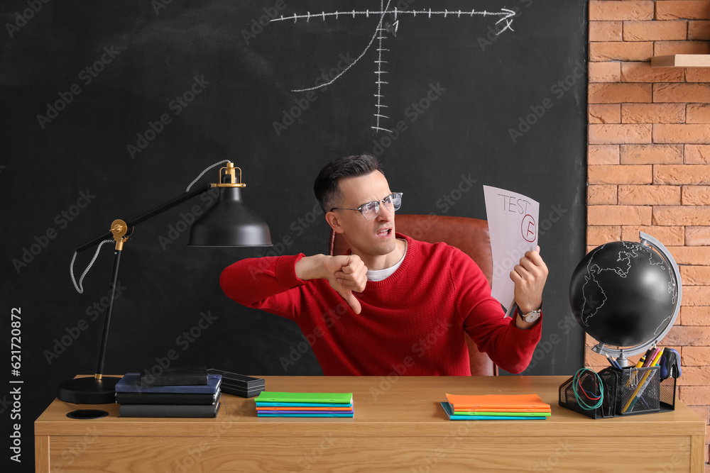 Male teacher with checked test sheet showing thumb-down at table in classroom