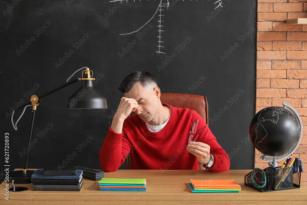 Tired male teacher sitting at table in classroom