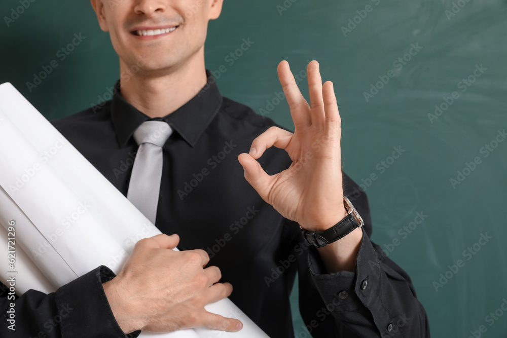 Male teacher with rolls of paper showing OK near chalkboard, closeup