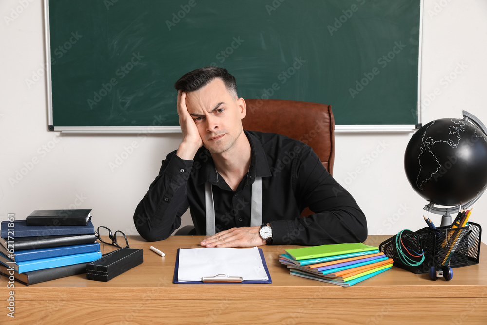 Tired male teacher sitting at table in classroom