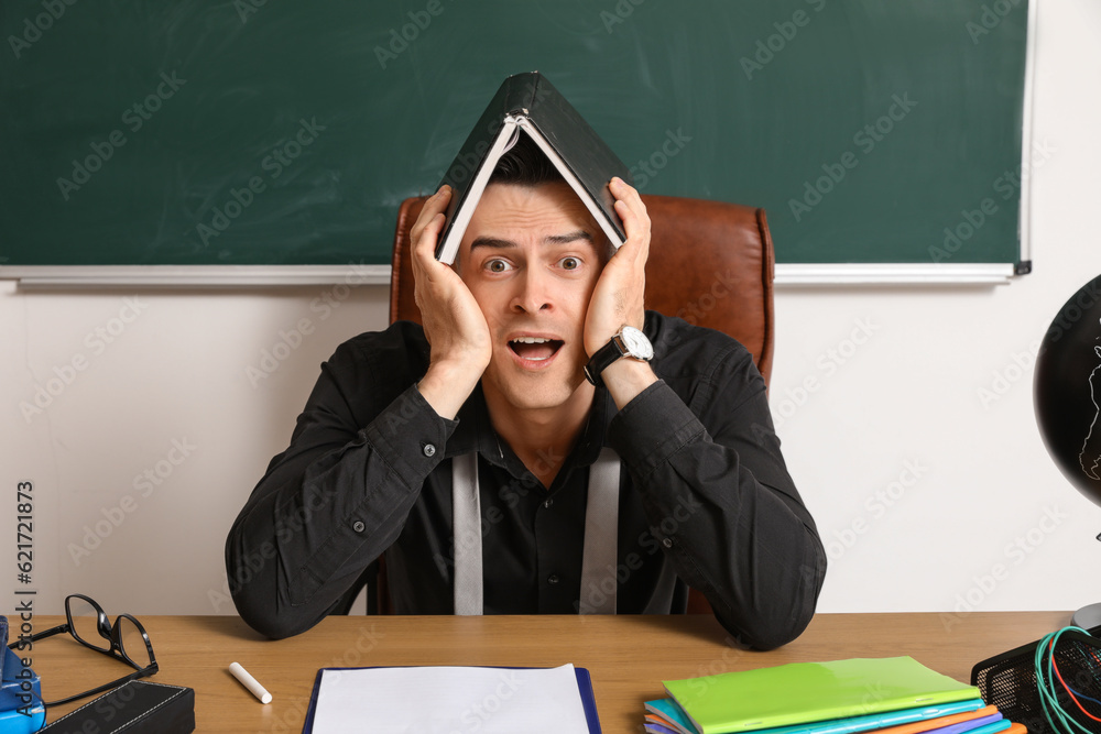 Stressed male teacher with book sitting at table in classroom