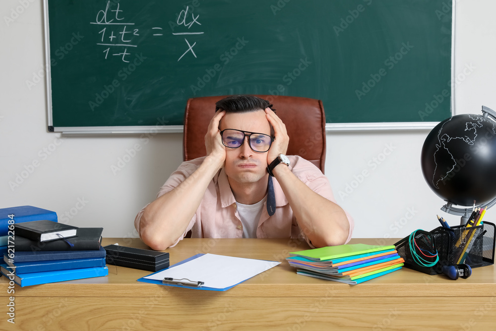 Stressed male teacher sitting at table in classroom