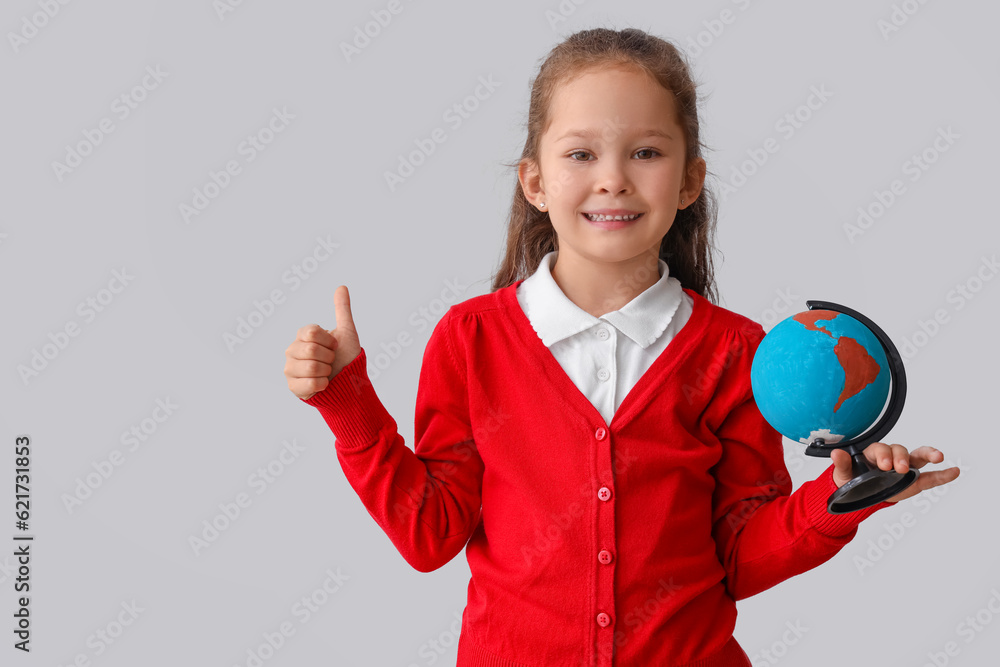 Little schoolgirl with globe showing thumb-up on grey background