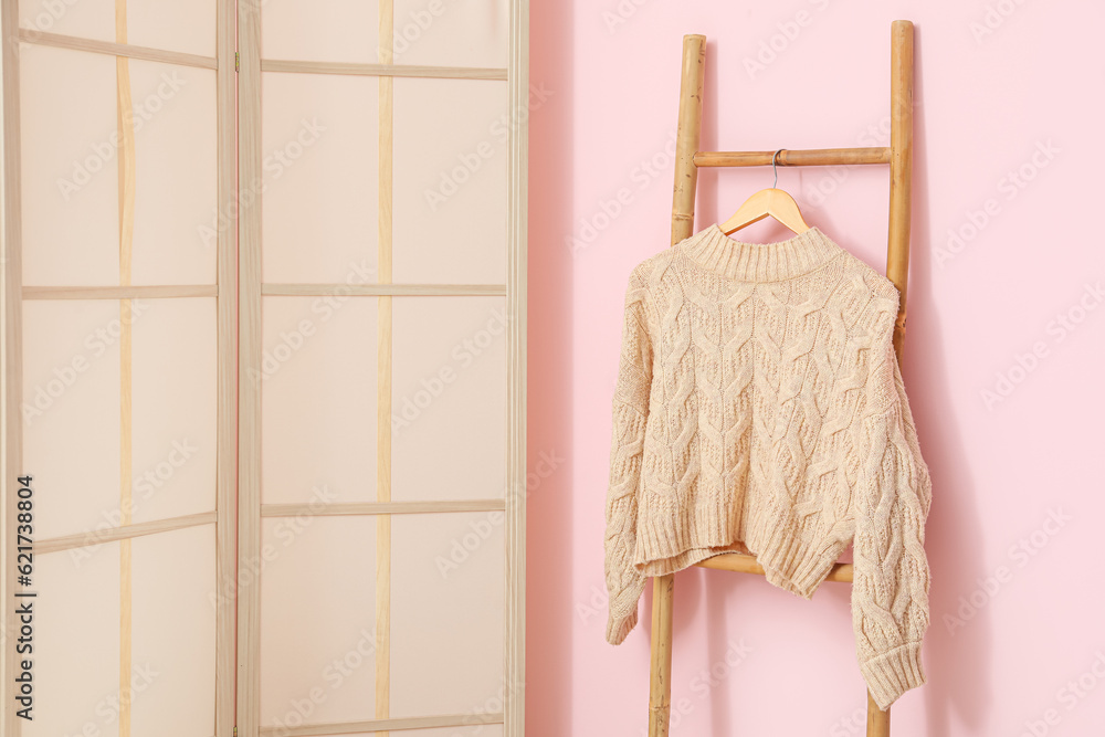 Sweater hanging on wooden ladder and folding screen near pink wall in room