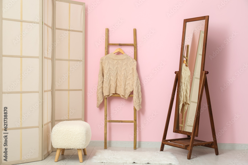 Sweater hanging on wooden ladder, folding screen, bench and mirror near pink wall in room