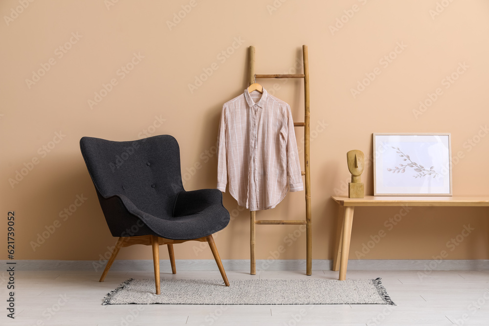 Shirt hanging on wooden ladder, armchair, table and picture near beige wall in room