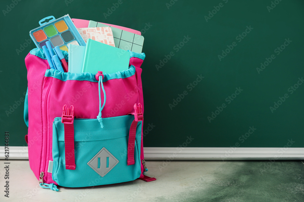 Colorful school backpack with notebooks, watercolors and pens on table near green chalkboard