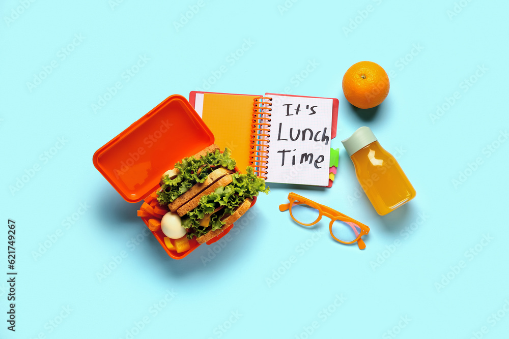 Notebook with text ITS LUNCH TIME, eyeglasses and tasty food in lunchbox on blue background