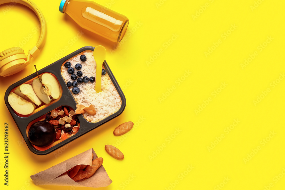 Bottle of juice, headphones, crackers and lunchbox with tasty food on yellow background