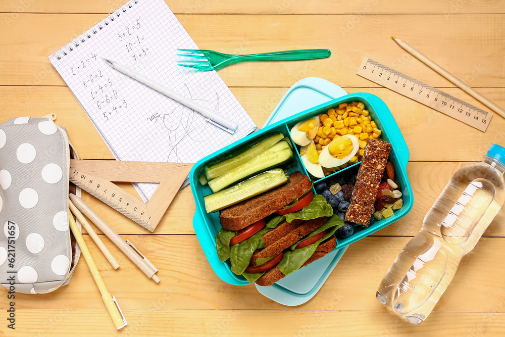 Bottle of water, different stationery and lunchbox with tasty food on brown wooden background