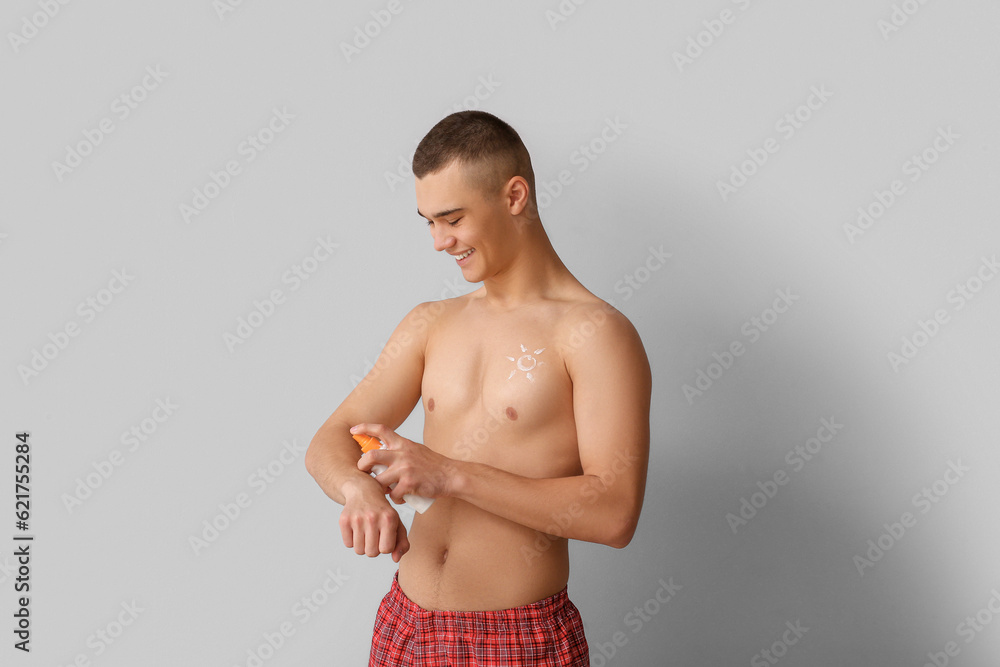 Happy young man applying sunscreen cream on his body against grey background