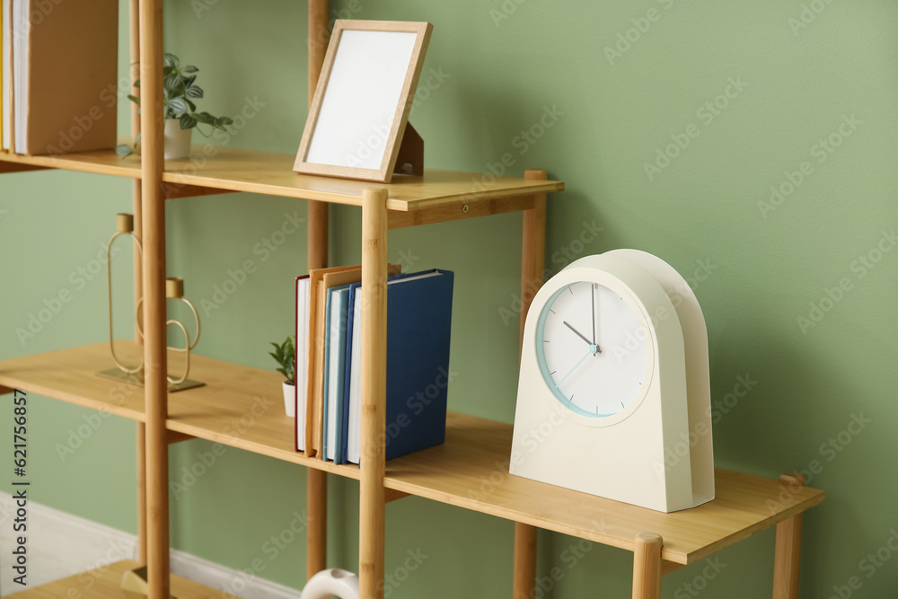 Shelving unit with books, clock and blank frame near green wall in room, closeup