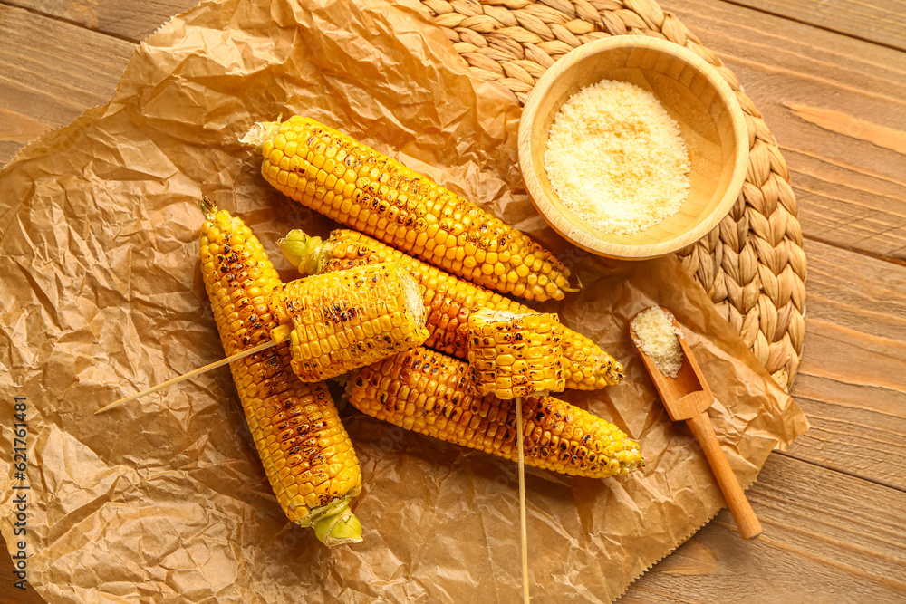 Baking paper with tasty grilled corn cobs and bowl of cheese on wooden background