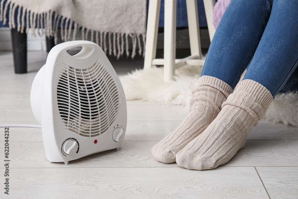 Woman in warm socks near electric fan heater at home, closeup